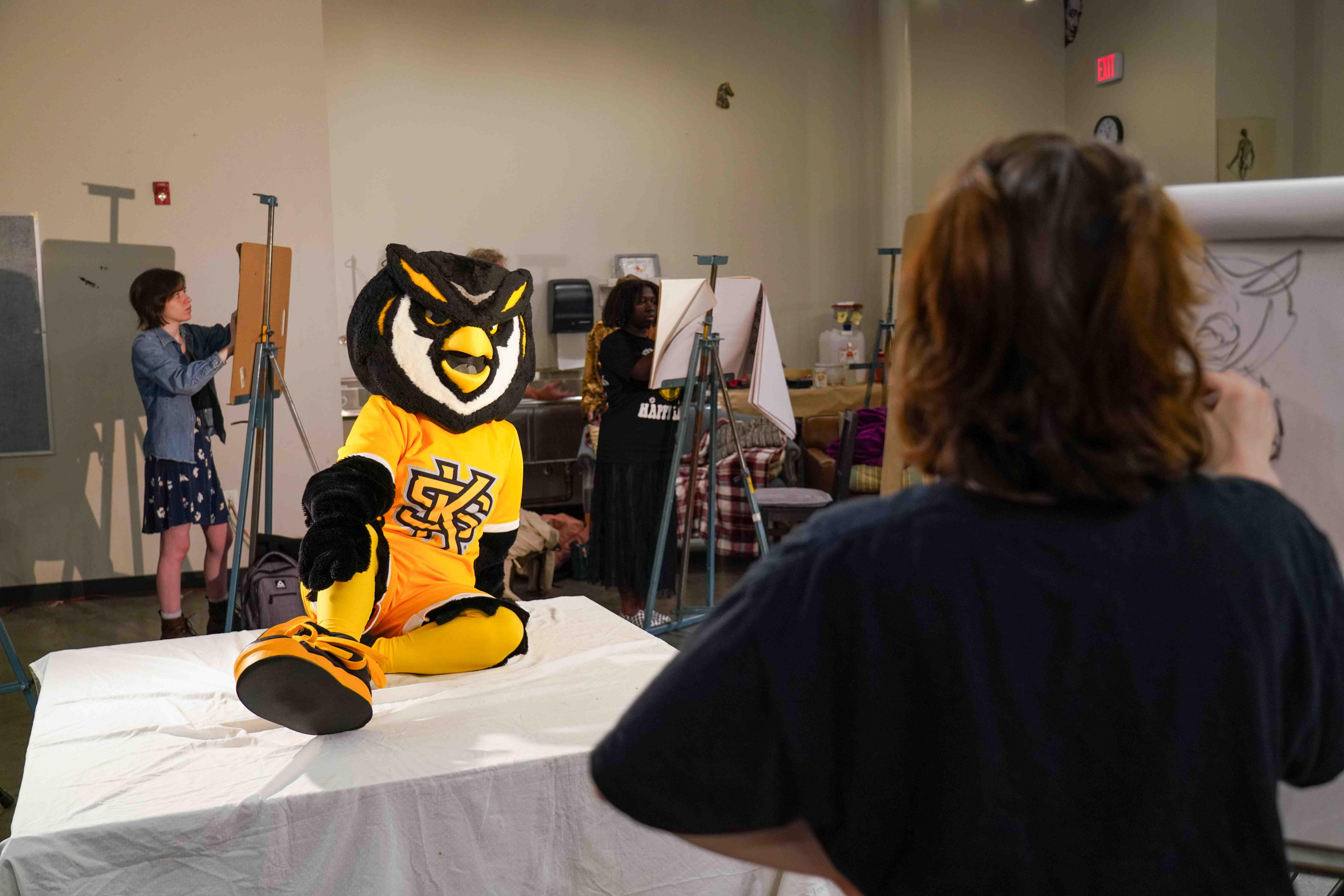 Scrappy as the model for a drawing class. / A costumed owl mascot seated on a platform in a classroom while students sketch on easels.