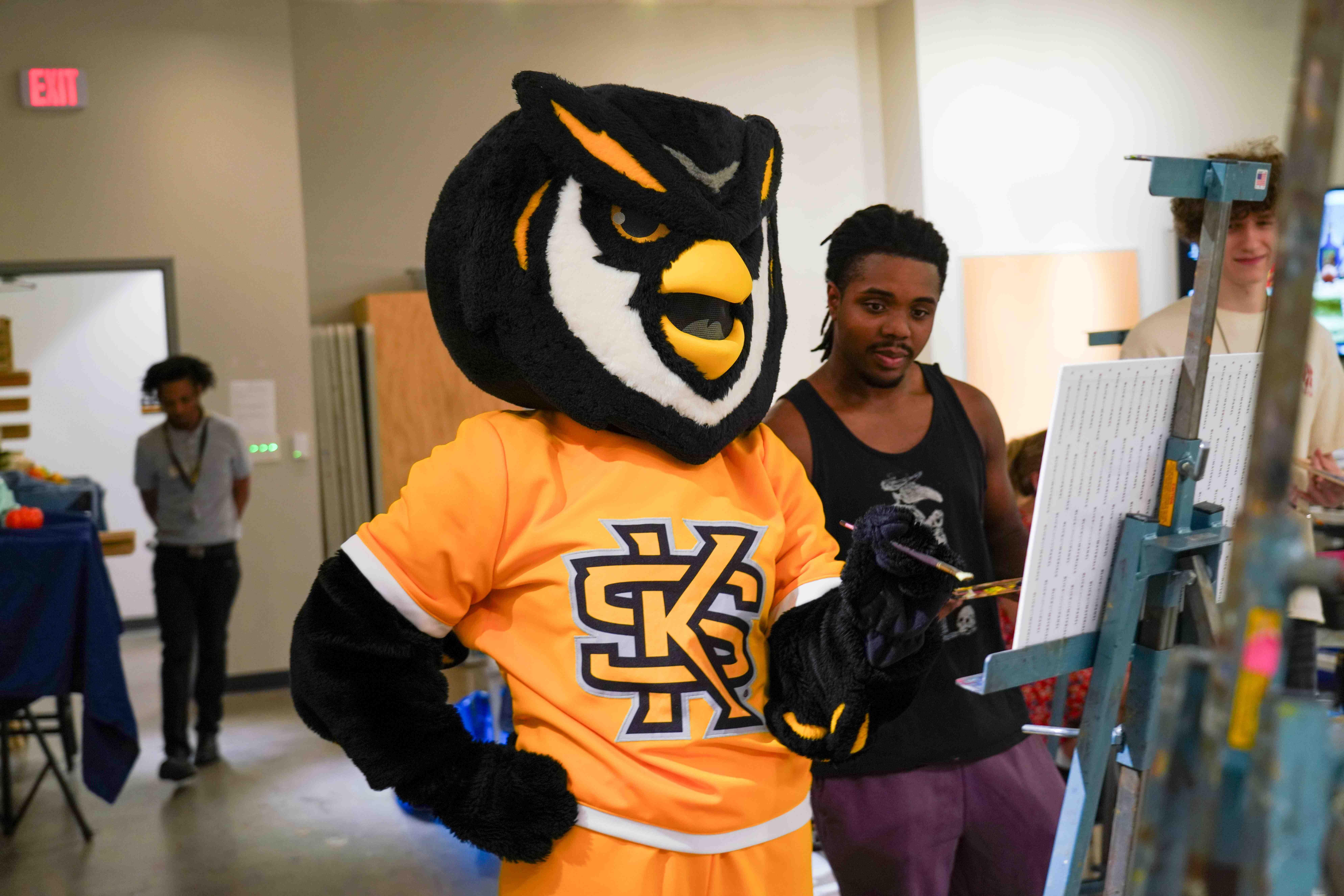 Scrappy visiting a painting class / A costumed owl mascot standing near easels in an art classroom while a person paints nearby.