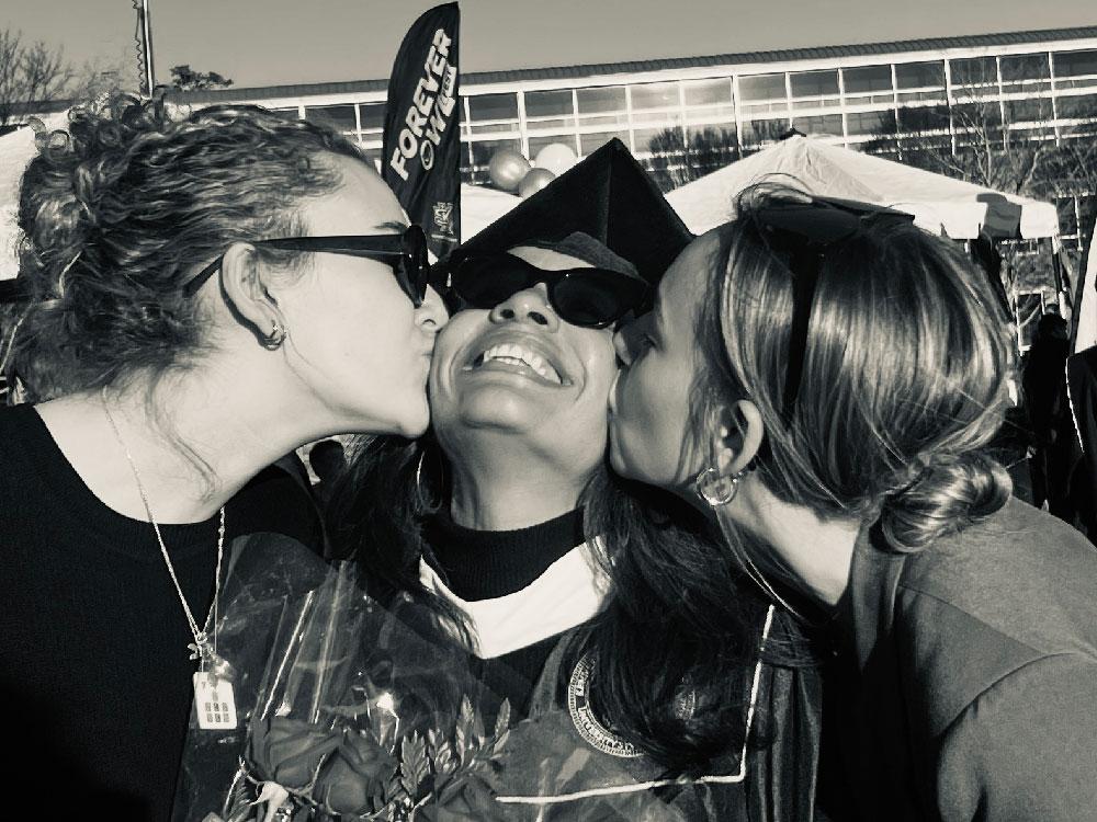 Patsy Rausch celebrates graduation / A person wearing a graduation cap and gown standing outdoors, holding a diploma cover while others stand nearby.