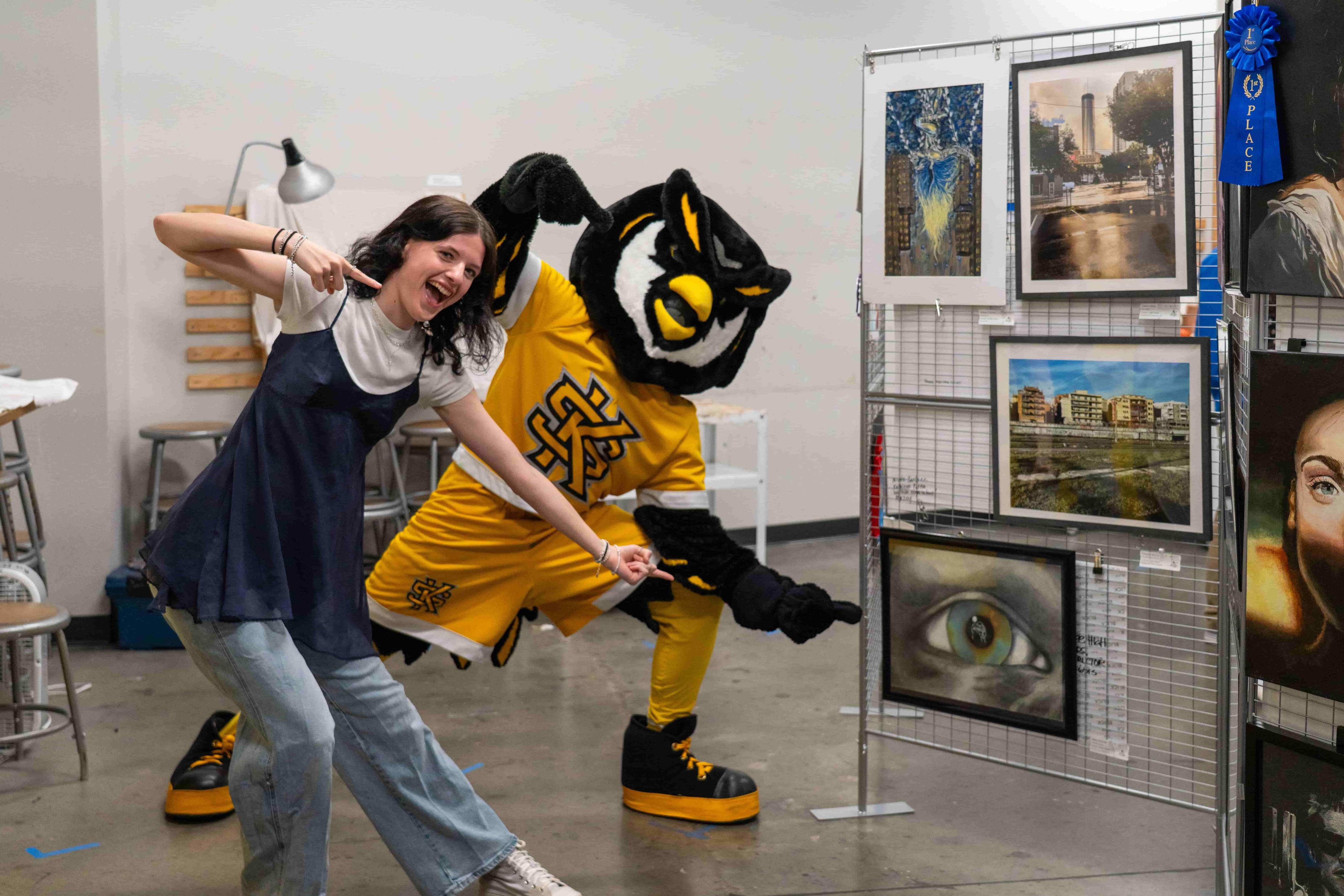 KSU mascot Scrappy the Owl with a K-12 student pointing at her work on display in the Art Show.   / 