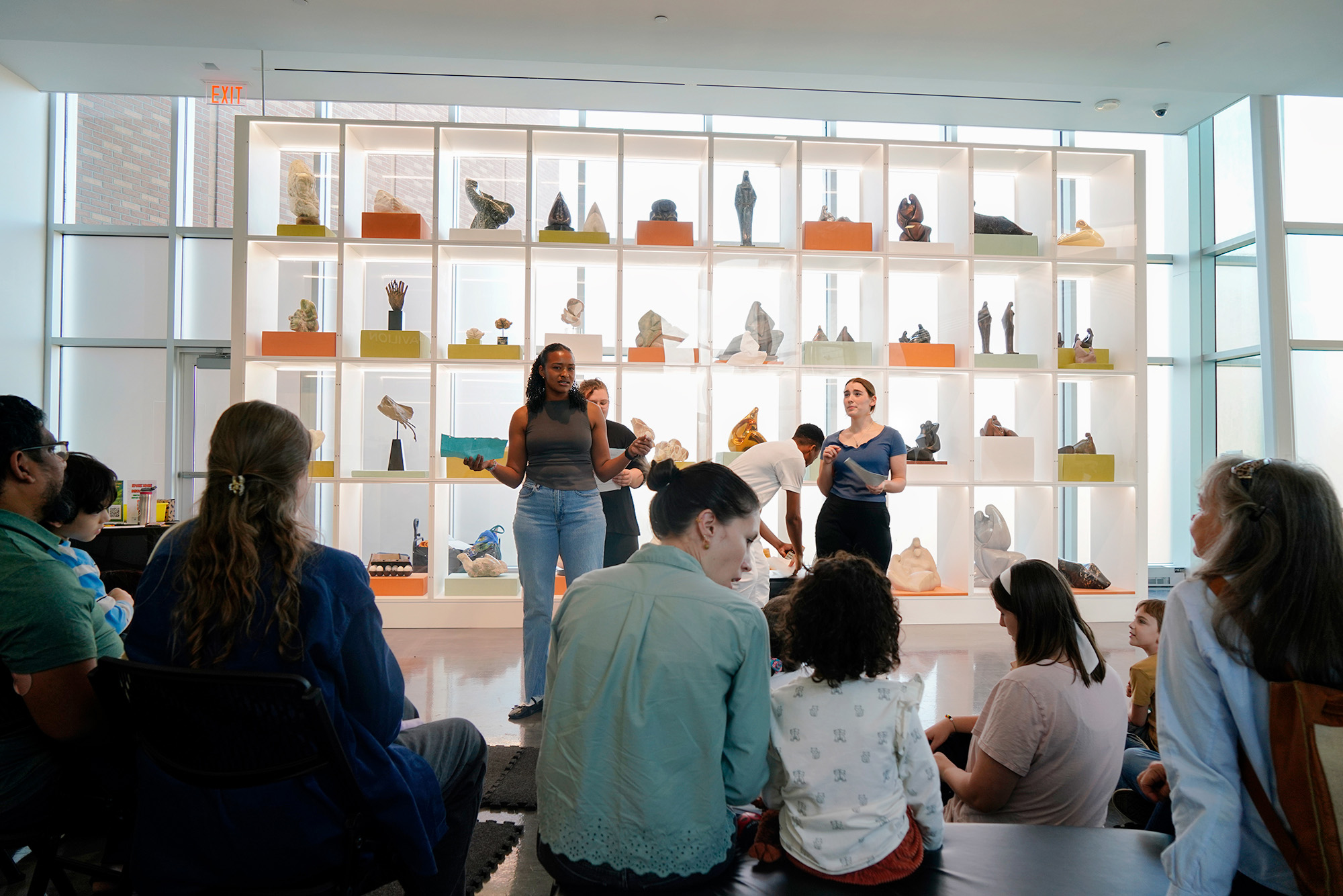 a group of onlookers watch as two women perform in front of a large grid full of sculptures