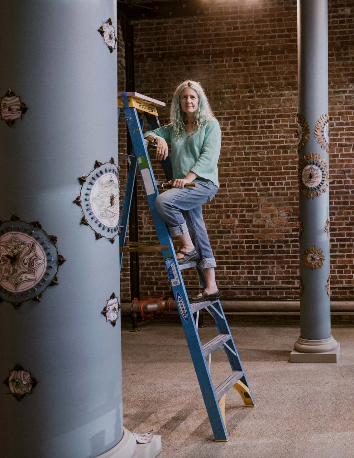 older woman on a ladder beside artwork on a column made of bugs
