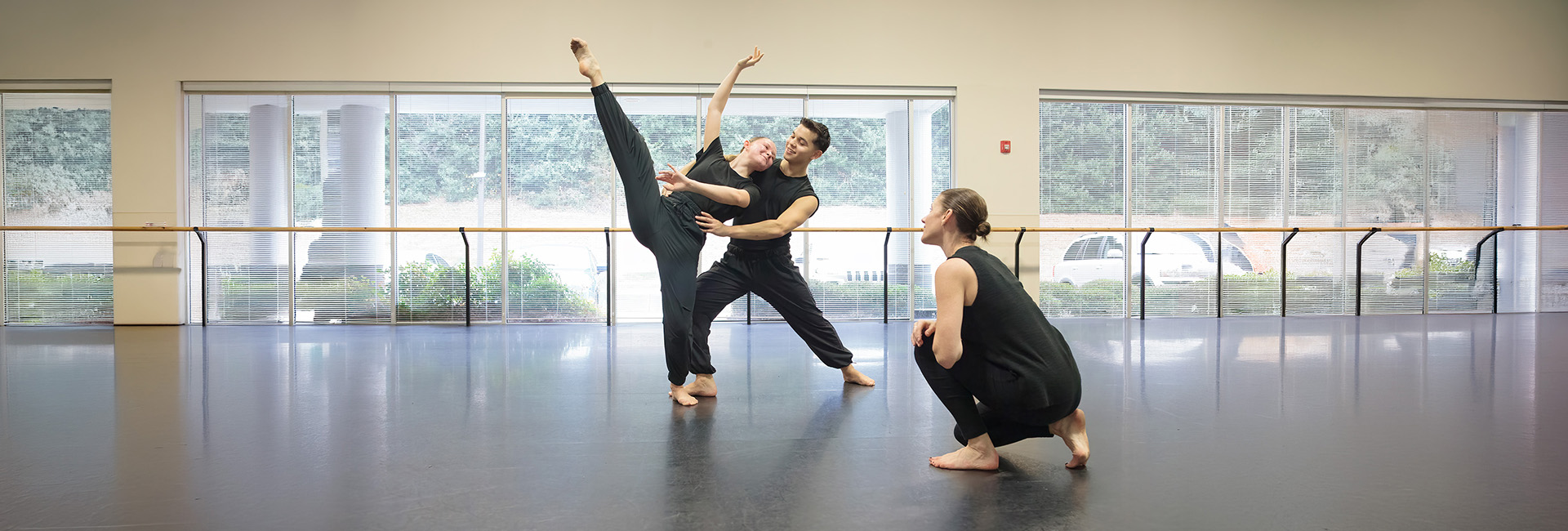 Kim Kenney 2025 KSU Dance Class Three dancers in black attire rehearse in a studio; two stand performing a lift while the third crouches nearby, observing. Large windows reveal greenery outside. 