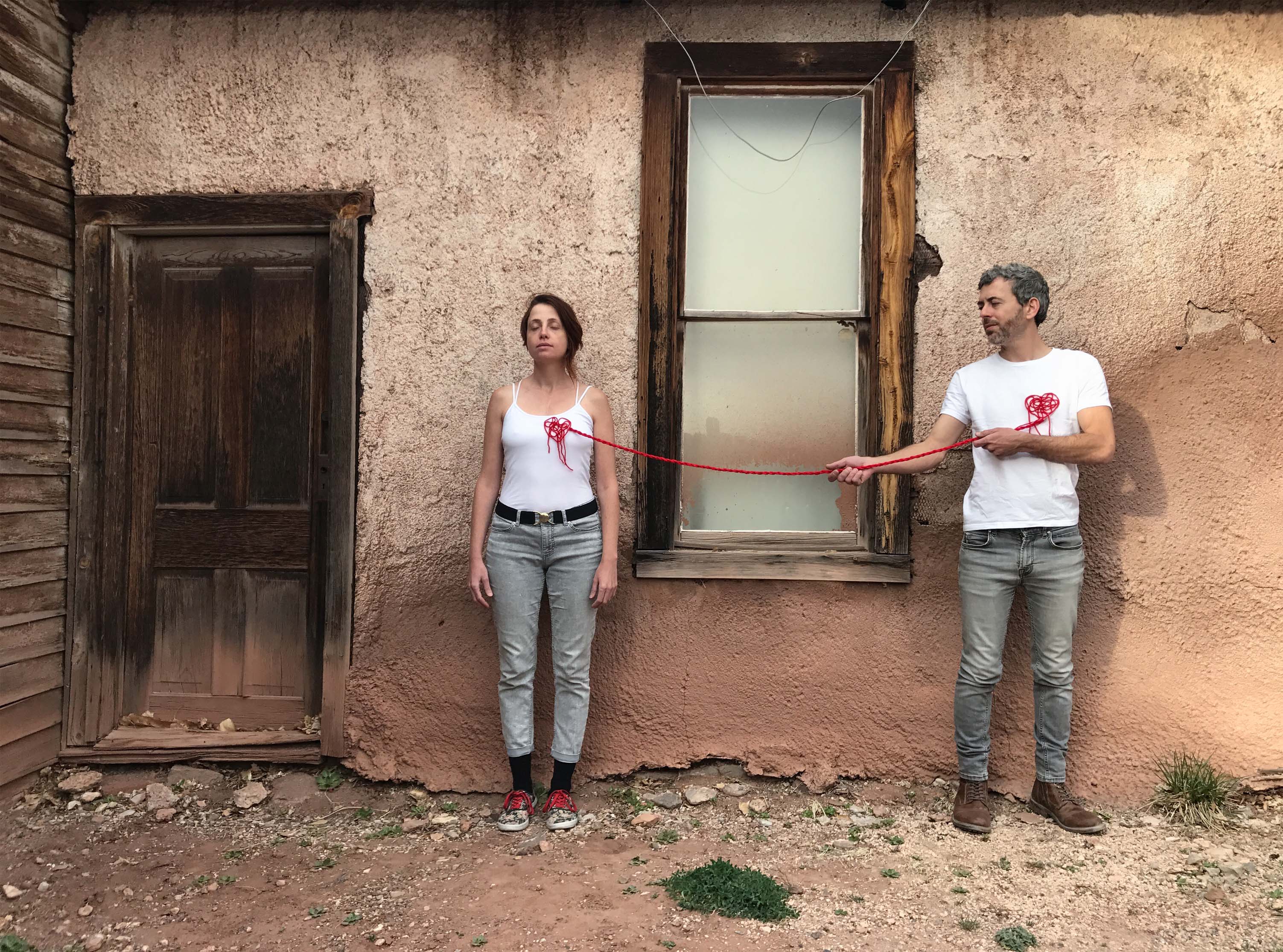 photograph of artists Maya Gelfman and Roie Avidan in front of wall with white shirts, red thread creating hearts on their chests and red string connecting their hearts 