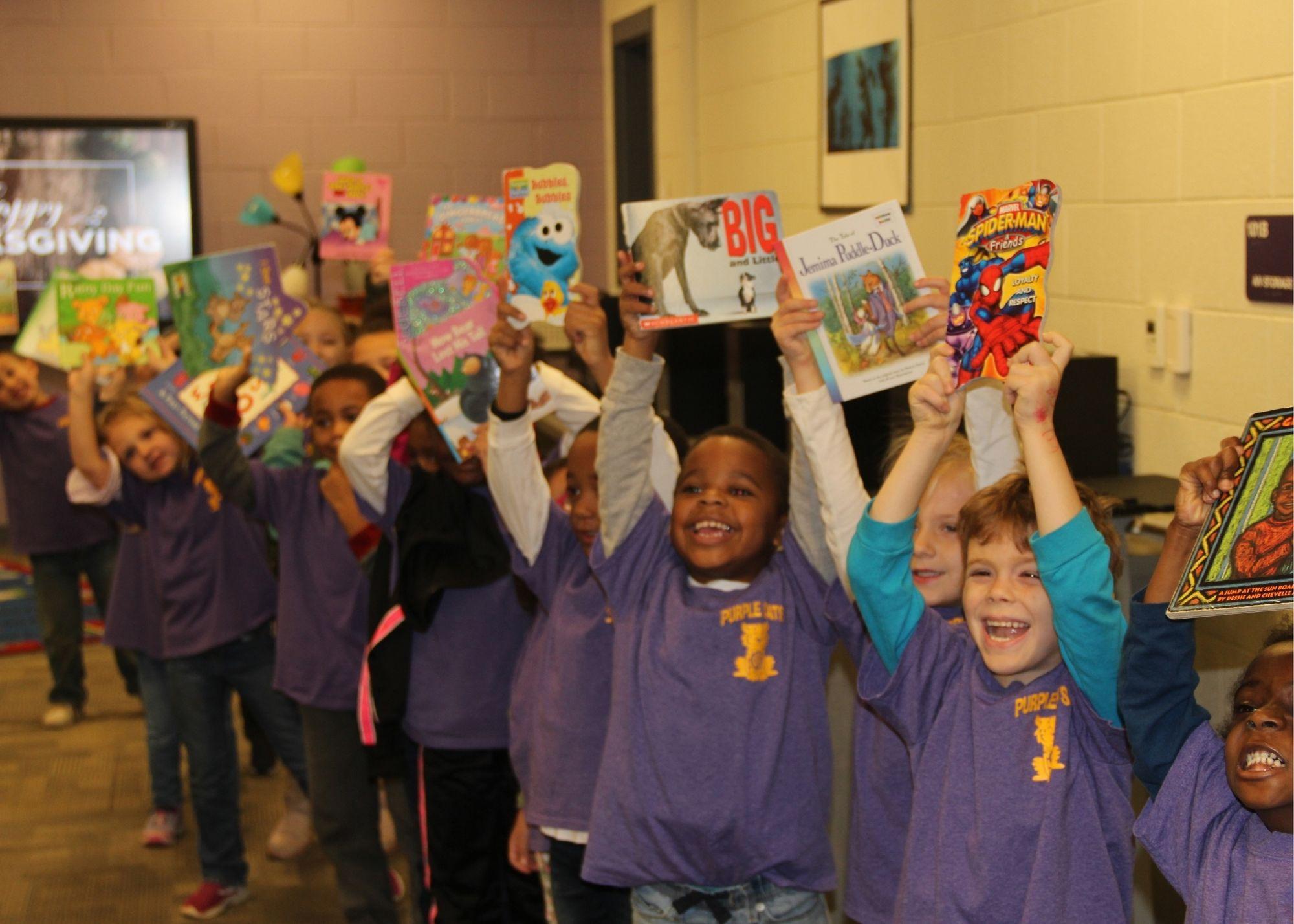 Students dressed in purple shirts smiling and holding books in the air.  / Students and staff from the Kennesaw State University and the Georgia Highlands College Paulding Site provided a special Book Drive for Villa Rica students. The groups went to Villa Rica Elementary School with hundreds of free books in order to give students access to a variety of reading material to build their reading skills. 
