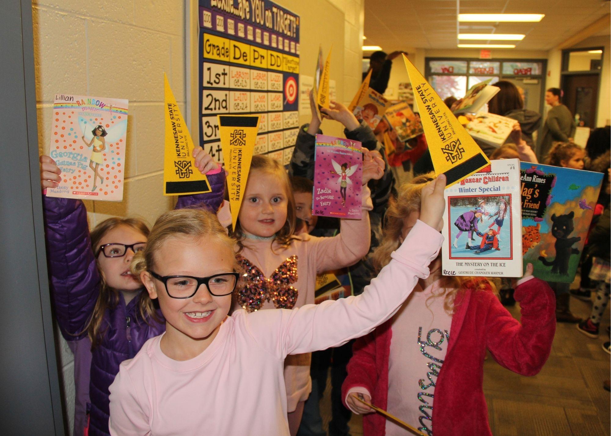 Young students in a hallway holding up books and school flags  / Students and staff from the Kennesaw State University and the Georgia Highlands College Paulding Site provided a special Book Drive for Villa Rica students. The groups went to Villa Rica Elementary School with hundreds of free books in order to give students access to a variety of reading material to build their reading skills. 
