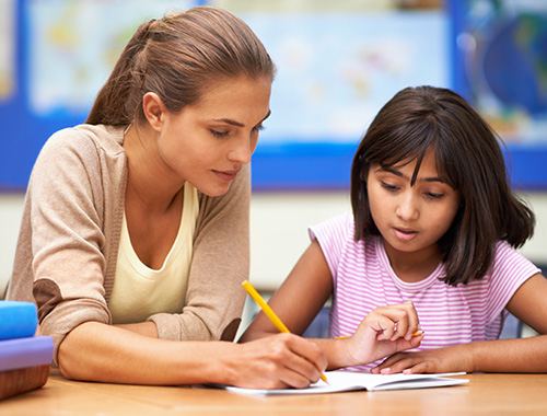 A teacher providing one-on-one assistance to a young student, guiding her through a writing exercise in a classroom setting.