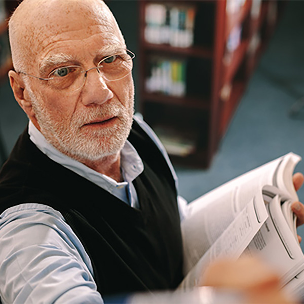 A professor with glasses and a white beard is standing in a library, holding an open book while writing on a whiteboard, symbolizing academic teaching and higher education.