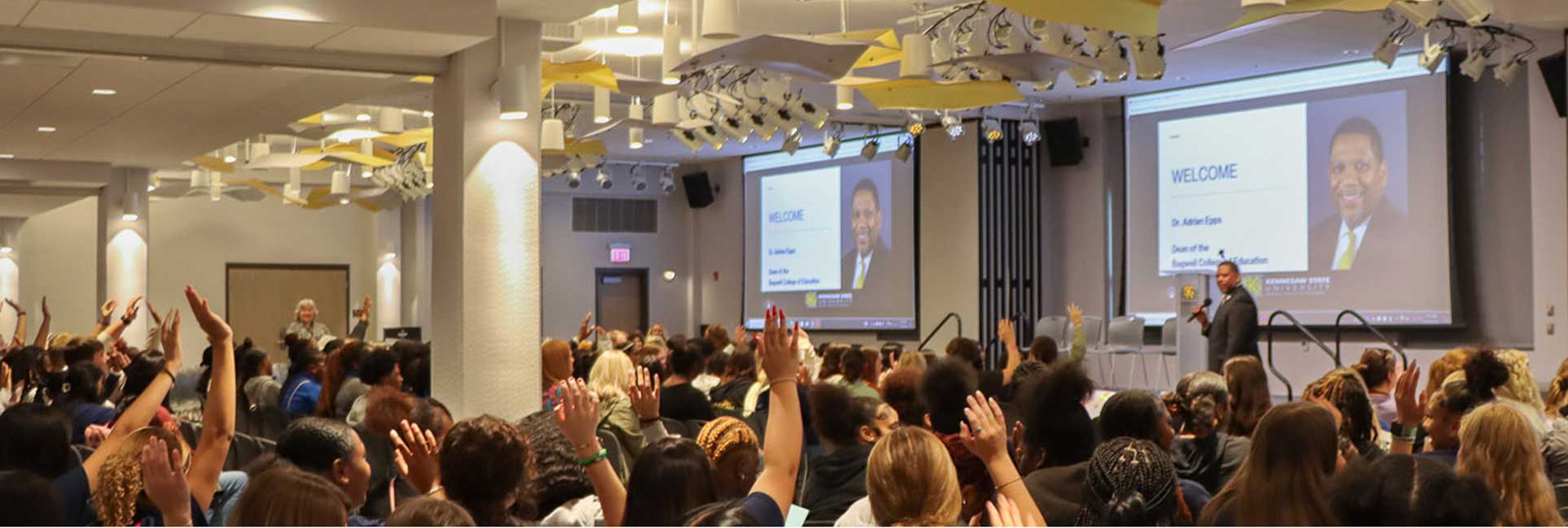 Photo of a large Aspiring Educators of Tomorrow Conference with attendees actively participating, raising their hands in engagement.