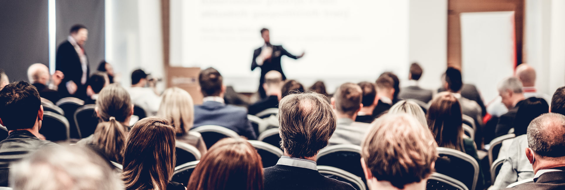 Audience members seated in a conference room, attentively listening to a speaker presenting at the front of the room.