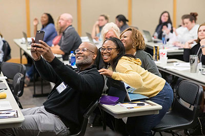 A diverse group of KSU students smiling and posing for a selfie in a brightly lit classroom setting.