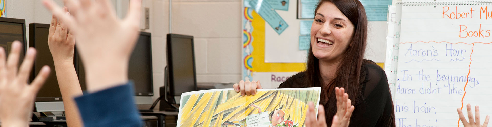 young adult reading a child's book to a group of young children raising their hand.