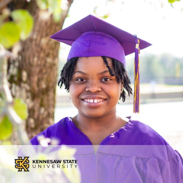 Anahjah Booker, a graduate in a purple cap and gown, smiling in front of a tree with the Kennesaw State University logo in the corner.
