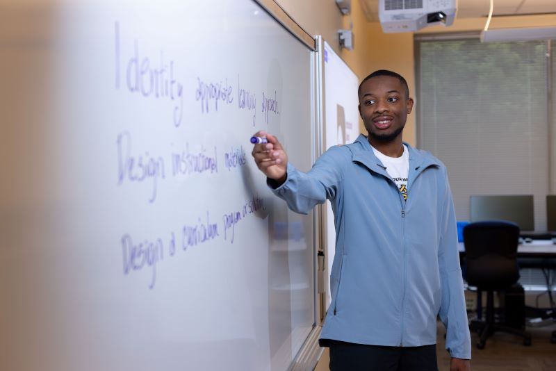 A student in a light blue jacket writes on a whiteboard in a classroom.
