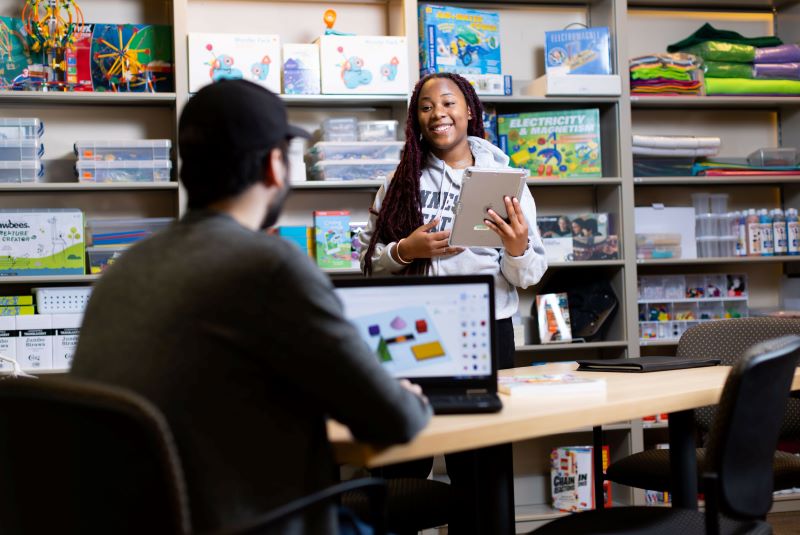 A woman stands while conversing with a man seated at a laptop in a classroom setting.
