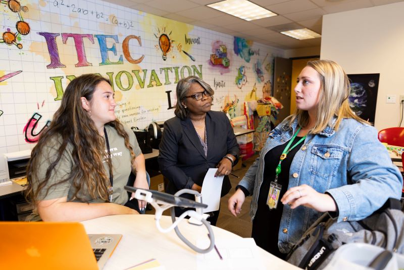 A group of three women engaged in conversation while one is seated in front of a laptop and the others are standing.