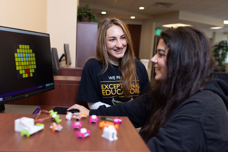 Two students smile while working on a computer and small robotics project.