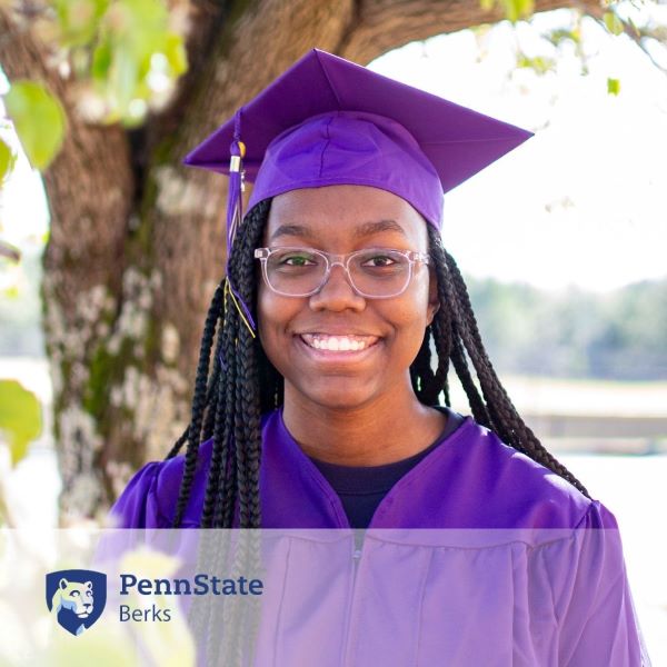 Janaya Allen, a graduate in a purple cap and gown, smiling in front of a tree with the Penn State Berks logo in the corner.