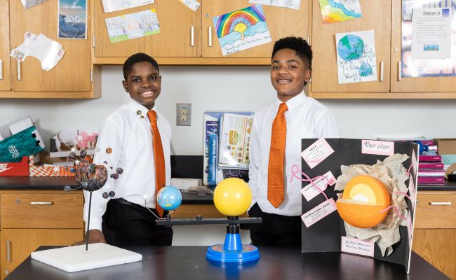 Two boys in white shirts with orange ties proudly display their science projects in front of them.