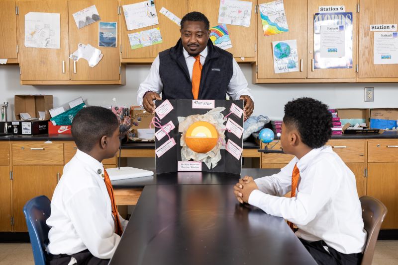 A teacher presents a solar system project to two students in a classroom.