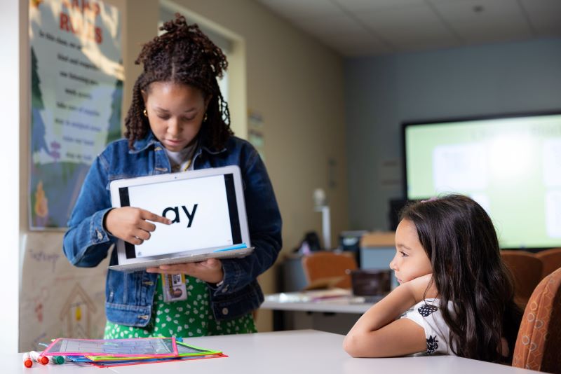 Teacher using a tablet to teach a young student phonics in a classroom.