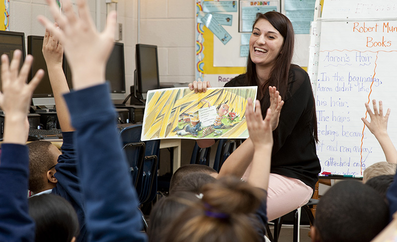 Teacher reading book showing pictures to students who are all raising their hands
