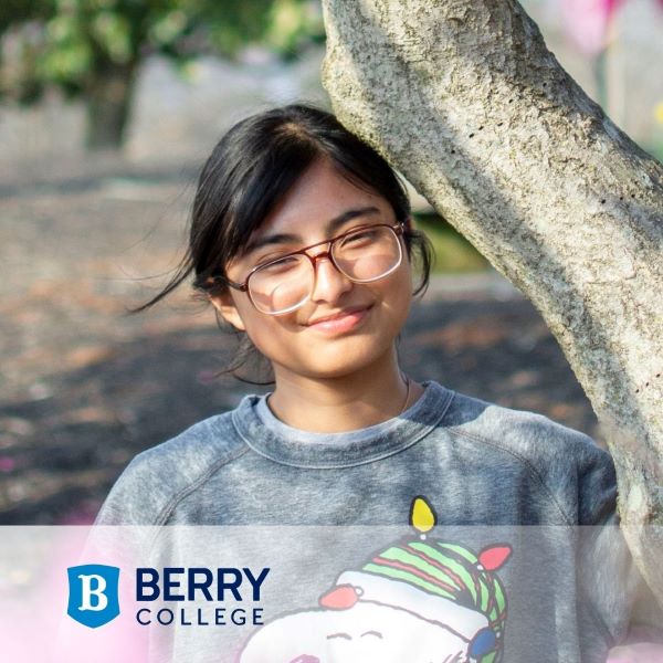 A student in a gray sweatshirt with a festive design, smiling while leaning against a tree. The Berry College logo is in the bottom left corner.