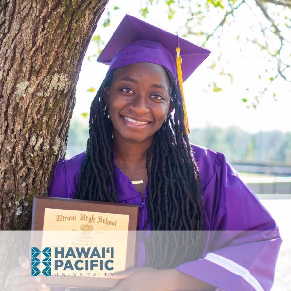 Ziphorah Tanis, a graduate in a purple cap and gown, smiling in front of a tree with the Hawaii Pacific University logo in the corner.