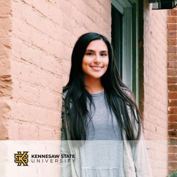 Emily Rodriguez Jacobo, smiling and leaning against a brick building, with the Kennesaw State University logo in the bottom left corner.