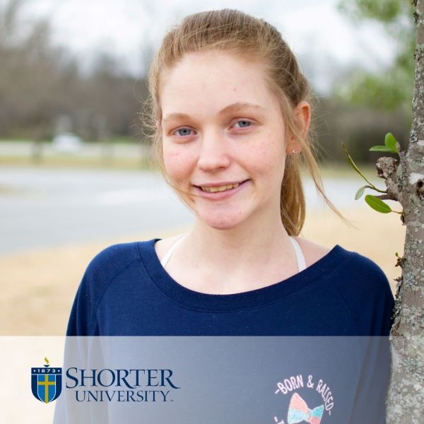 Elizabeth Shupp, smiling and leaning against a tree, with the Shorter University logo in the bottom left corner.