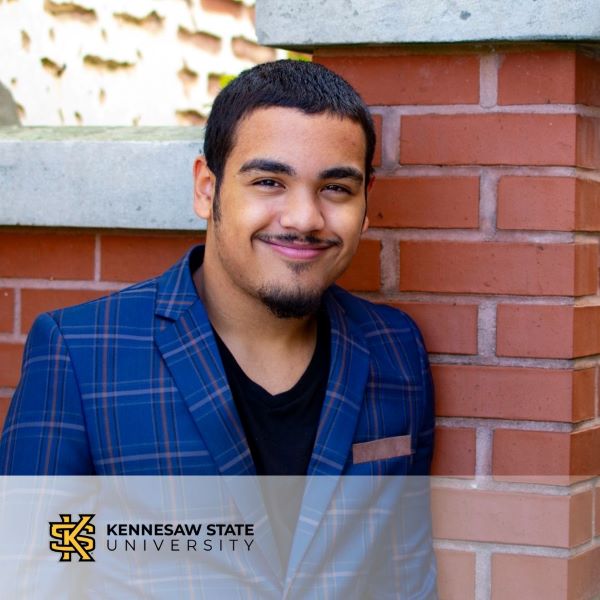 Elio Vargas, smiling and leaning against a brick wall, with the Kennesaw State University logo in the bottom left corner.