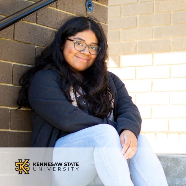 Sarai Perez, a smiling student leaning against a brick wall, with the Kennesaw State University logo in the bottom left corner.