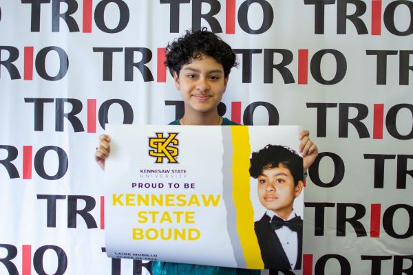 A student holds a sign that says they are "Kennesaw State Bound" in front of a backdrop with the word "TRIO" repeated.