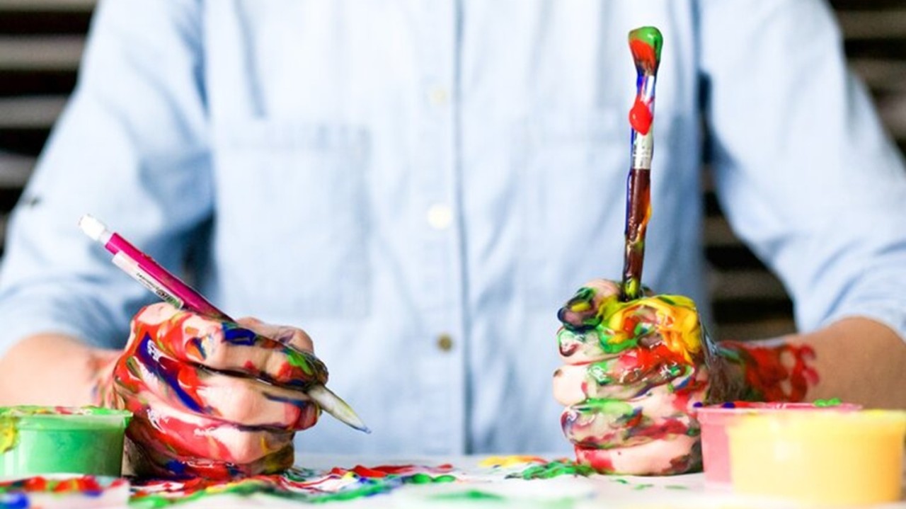 person in blue button up shirt with a pencil and paint brush in had covered in paint
