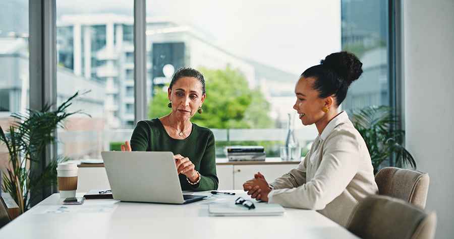 two women in a brightly lite office look over a laptop