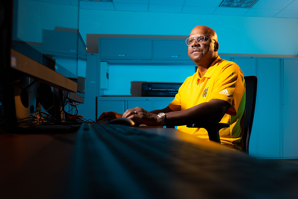 A man in a KSU-branded shirt sits at a computer, smiling