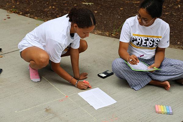 PRESS students from the college of science and mathematics sit on the pavement, writing math equations with chalk as part of an engaging group activity. / PRESS students from the college of science and mathematics sit on the pavement, writing math equations with chalk as part of an engaging group activity.