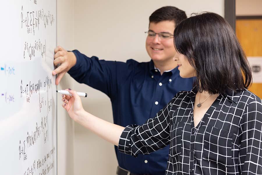Research student writing on a white board working on formulas with another student on a research project. / Research student writing on a white board working on formulas with another student on a research project.
