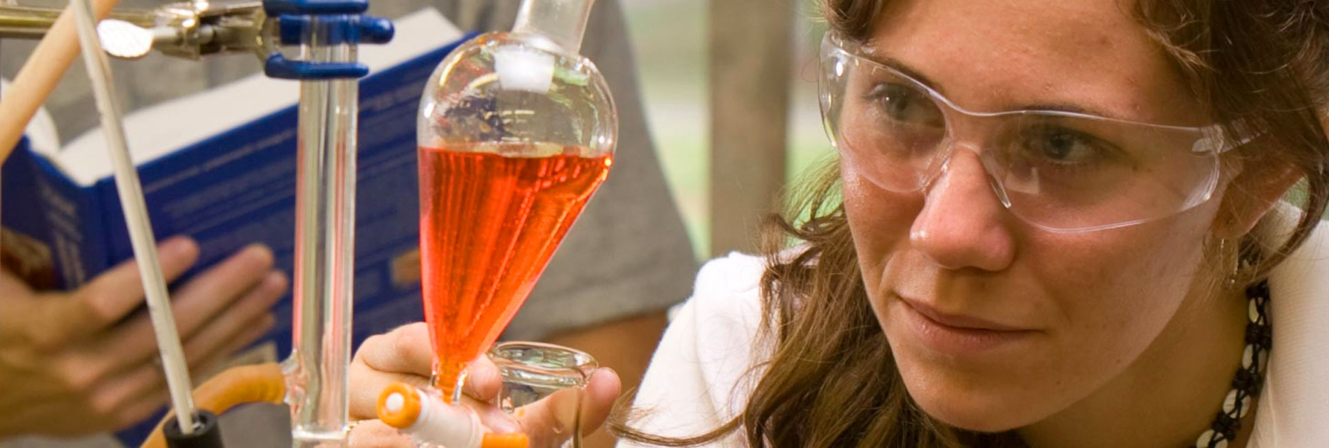 A focused Kennesaw State graduate student wearing protective safety goggles observes a laboratory experiment. She holds a flask containing a bright orange liquid and appears to be adjusting the equipment in a scientific setting, highlighting concentration and precision during the experiment.