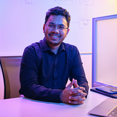 Abm adnan azmee in a dark shirt sits at a desk with a laptop, smiling confidently. The room is softly lit in blue and purple, creating a calm, professional atmosphere.