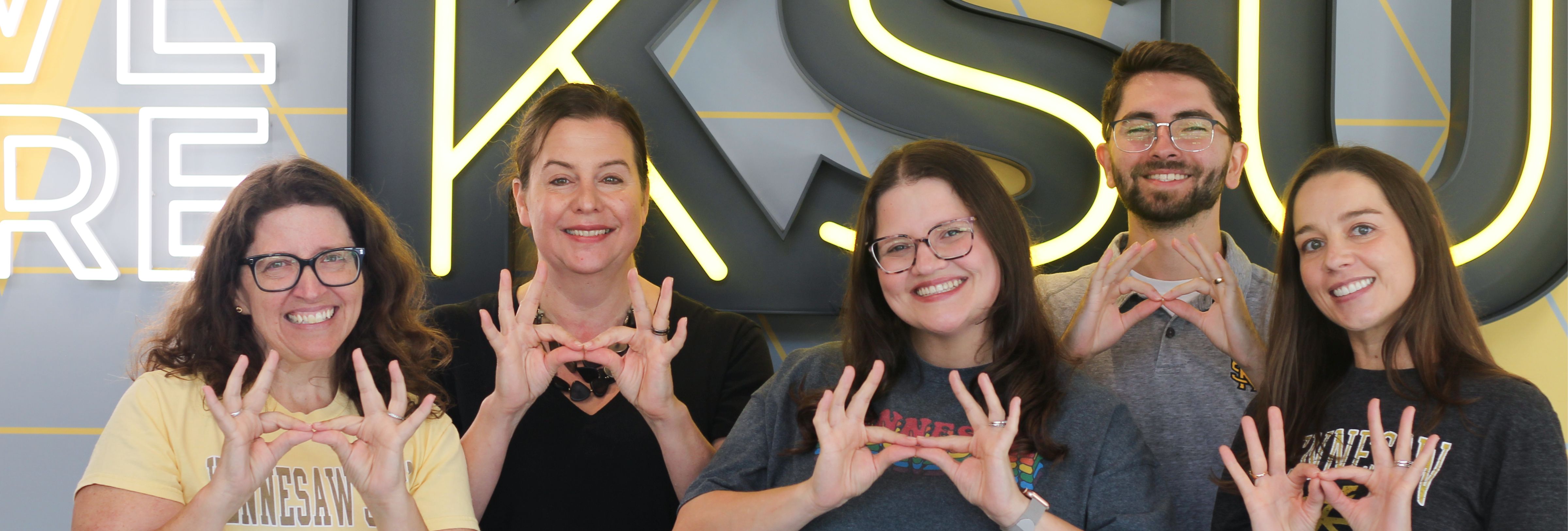 Group of Honors advisors smile and hold the "owl eyes" hand sign while standing in front of a We Are KSU sign.