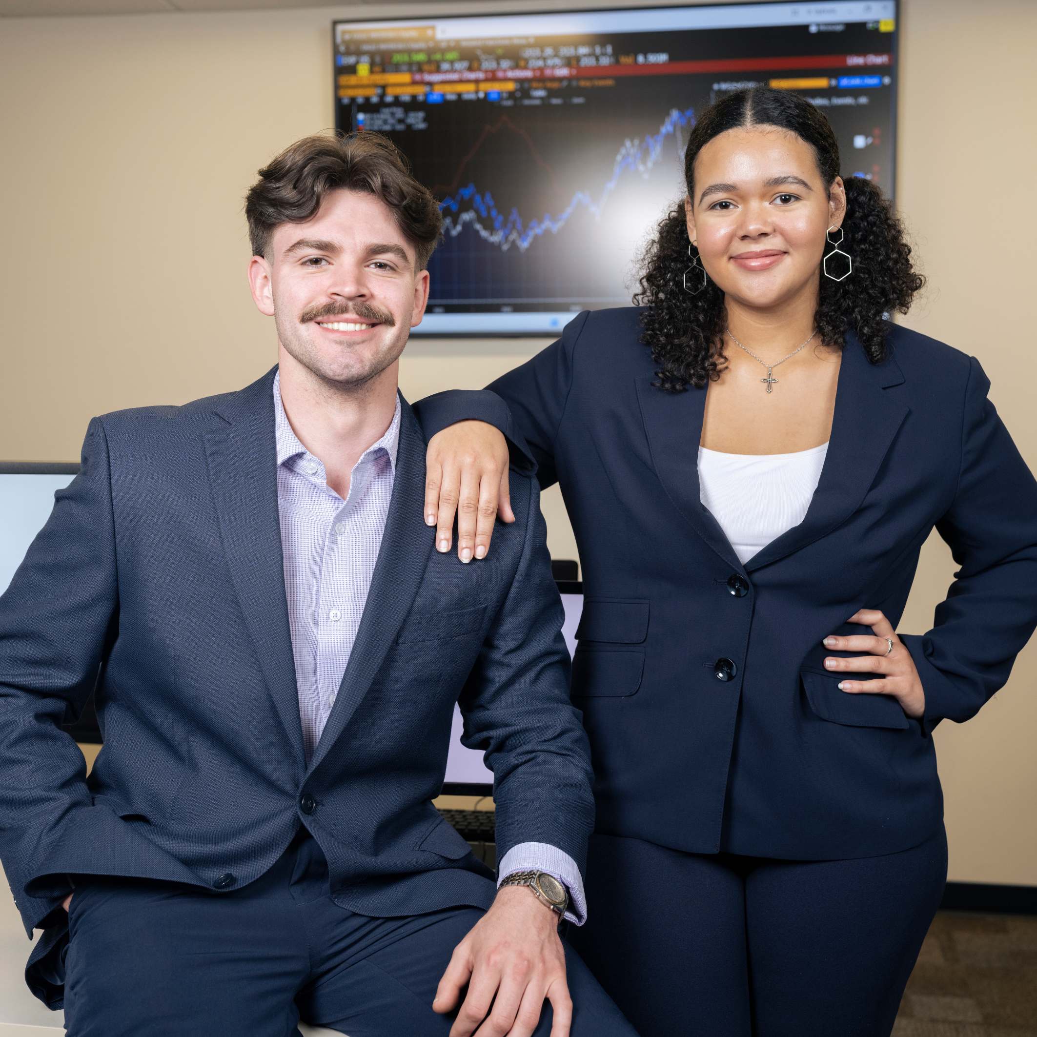 Two students in professional attire posing indoors, with a computer screen behind them showing financial data and stock charts.
