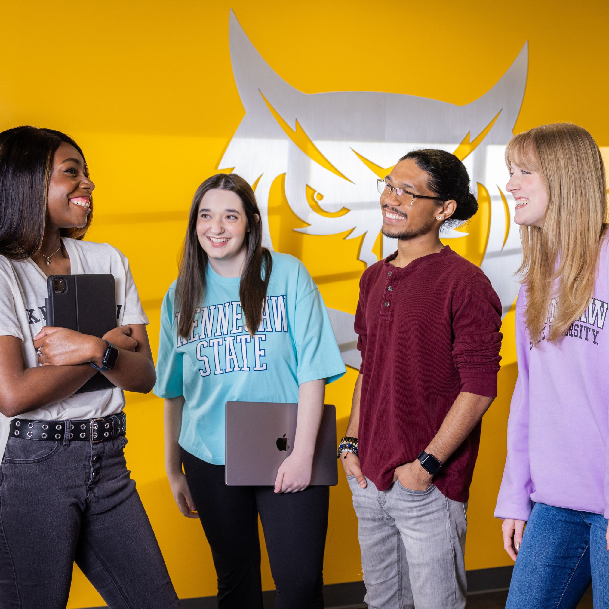 Four KSU students stand in a circle while wearing Kennesaw State University branded shirts.