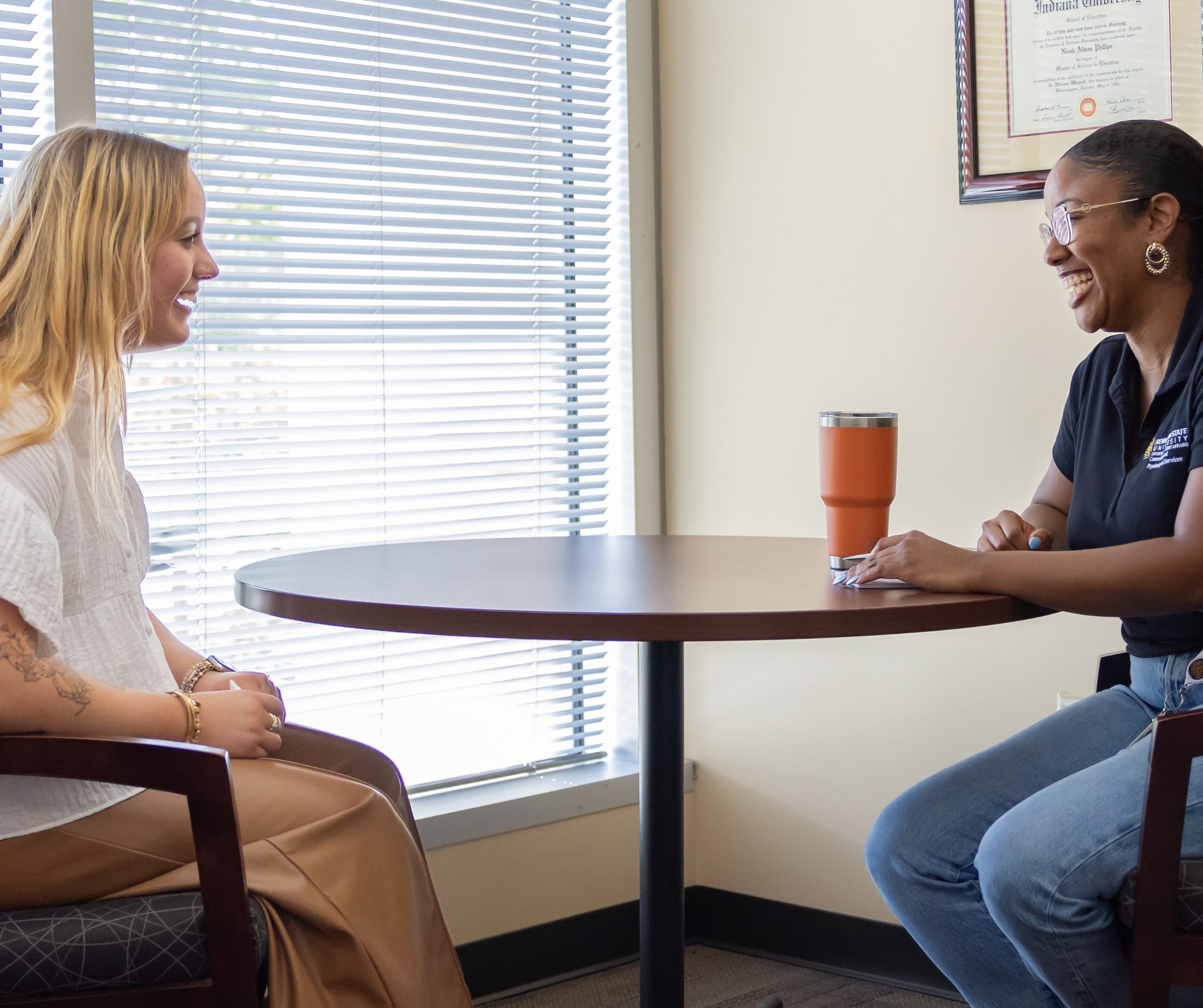 A student and staff member sit and talk at a table near a window.