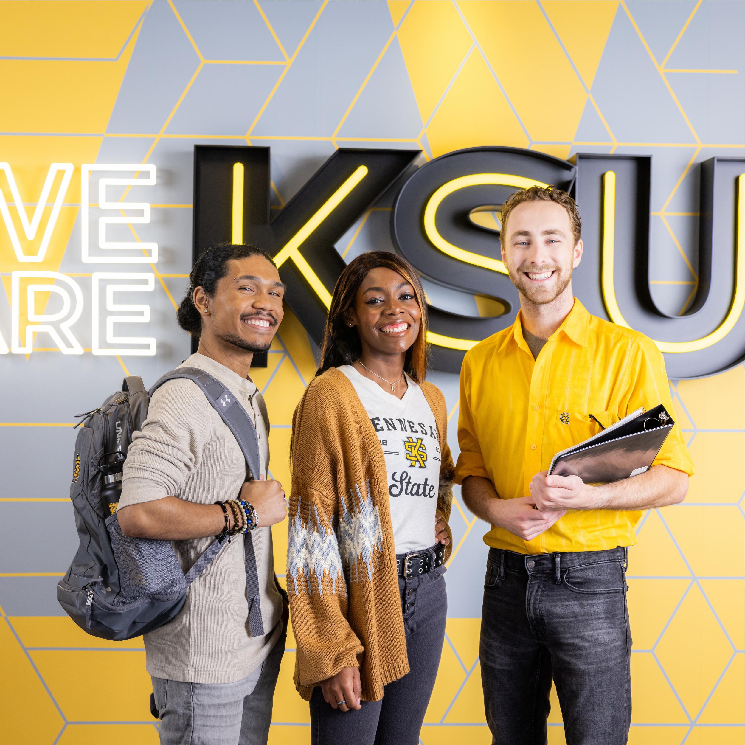 Three Kennesaw State University's smile in front of a sign that says "We Are KSU" while wearing black and yellow.