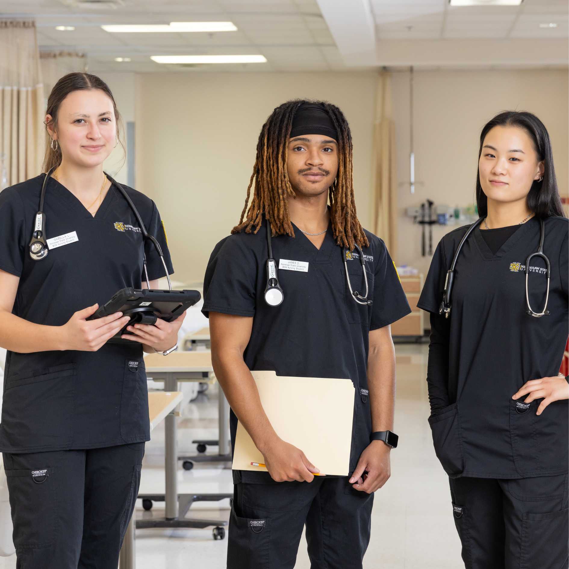 A group of three diverse students stand in a nursing lab with blacks scrubs on and equipment in the background..