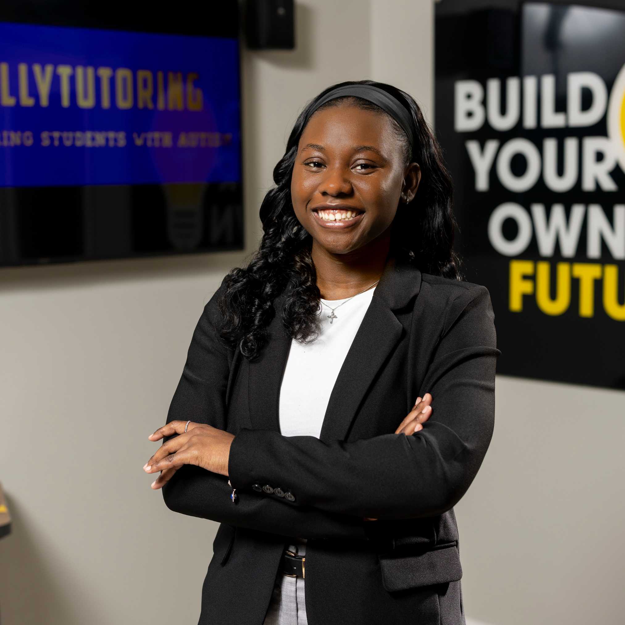 Young black female wearing a black blazer and smiles with arms folded.
