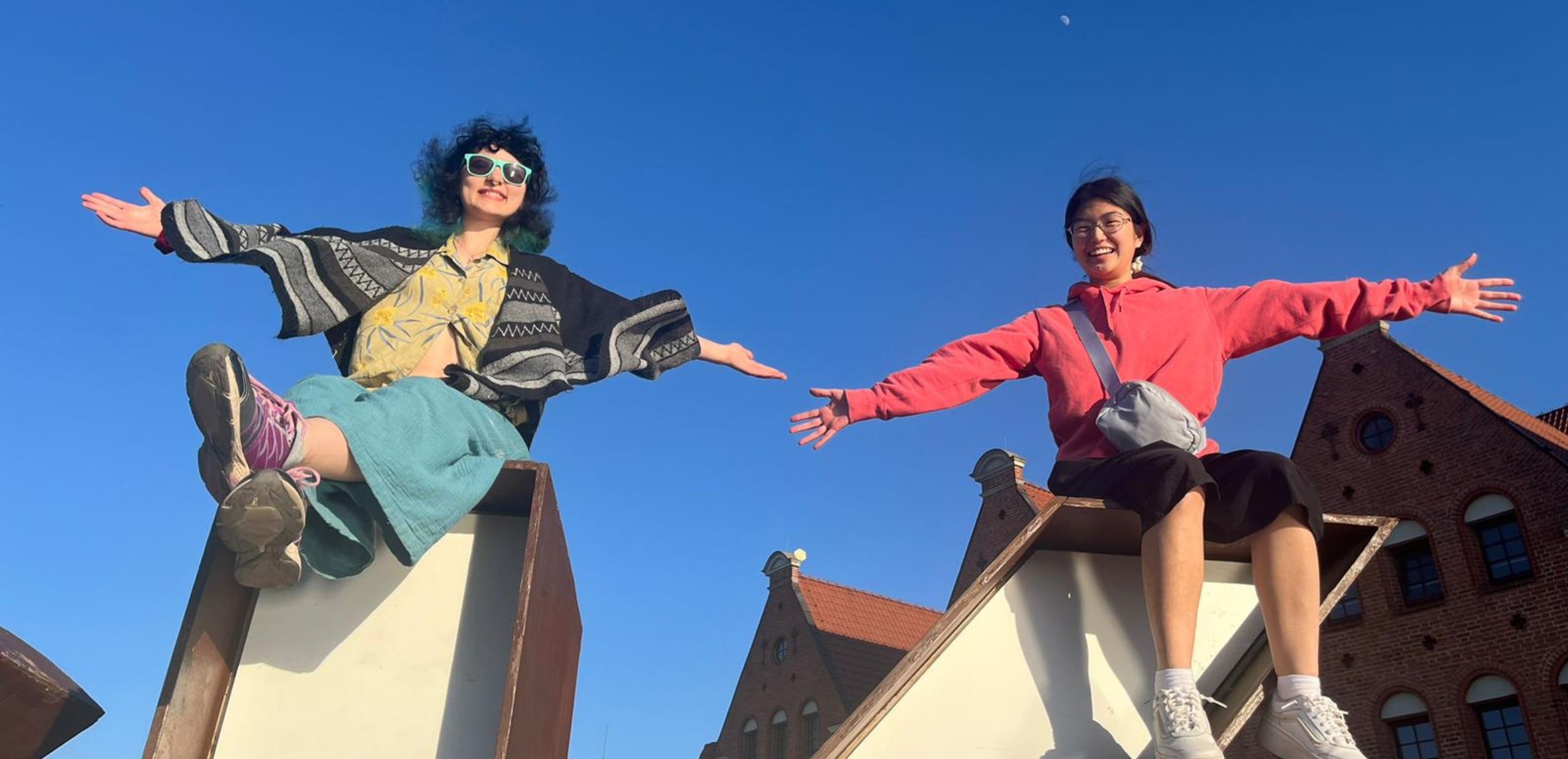 2 students smile and sit on top of a building with their arms outstretched
