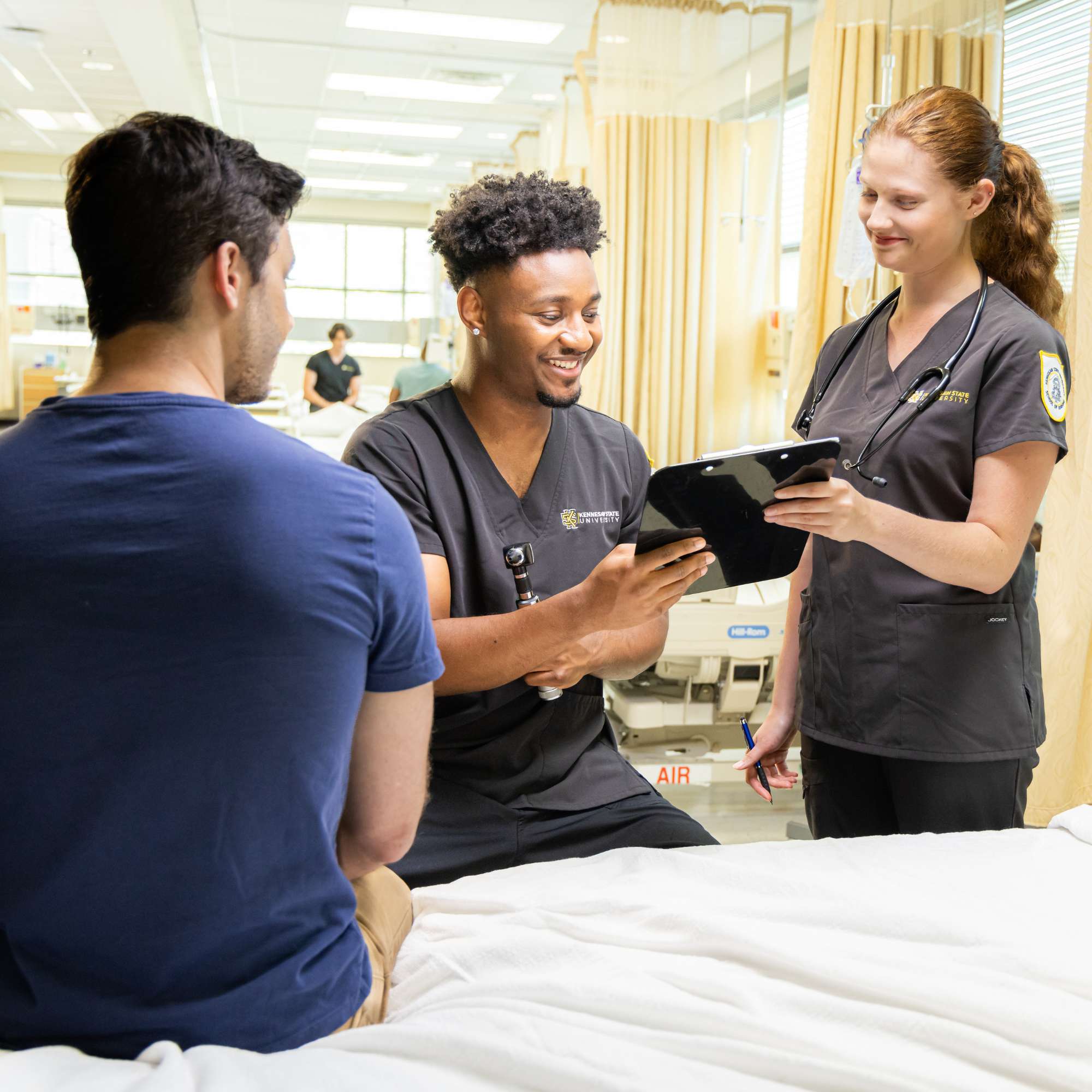 KSU nursing students in a nursing simulation lab review information on a tablet beside a hospital bed, with medical equipment and privacy curtains visible in the background.