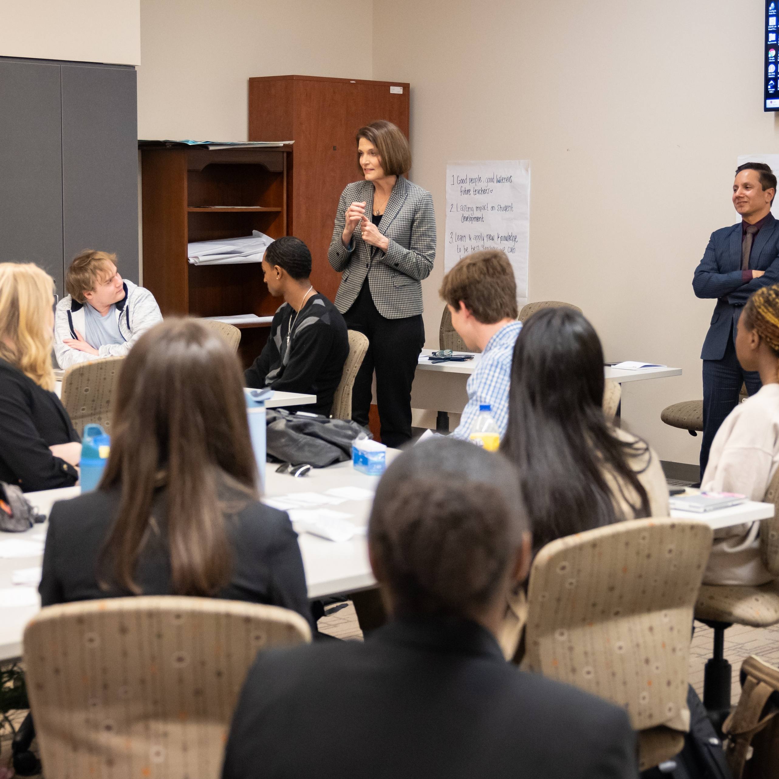 A classroom workshop is in progress with students seated around white tables arranged in a group setting. A speaker wearing a patterned blazer stands at the front of the room addressing participants, while another presenter stands to the side.